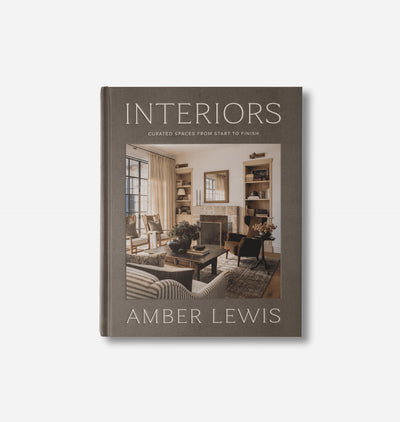 The book 'Interiors: Curated Spaces from Start to Finish' by Amber Lewis, featuring a dark taupe-gray cover with a central photograph of a cozy living room, set against a light gray background.