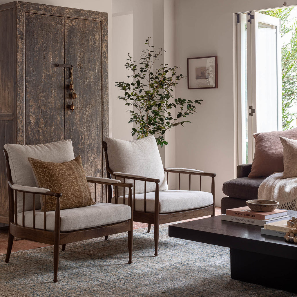 A styled living room featuring two wooden armchairs with off-white cushions and patterned pillows, a dark wood coffee table, a rustic wooden cabinet, and a potted plant.