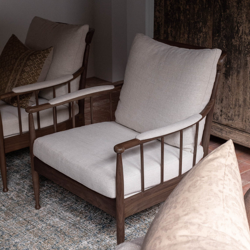 A wooden armchair with light beige linen cushions sits in a styled interior setting with a patterned rug, rustic wooden wall paneling, and another matching chair in the background.