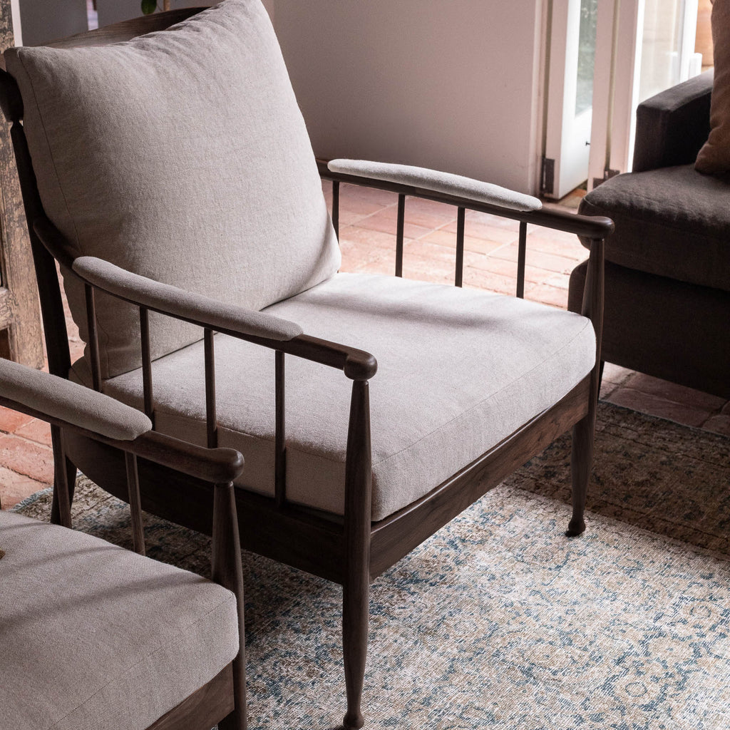 A mid-century modern armchair with a dark wood frame and light beige textured fabric cushions, shown in a styled living room setting on a patterned rug.