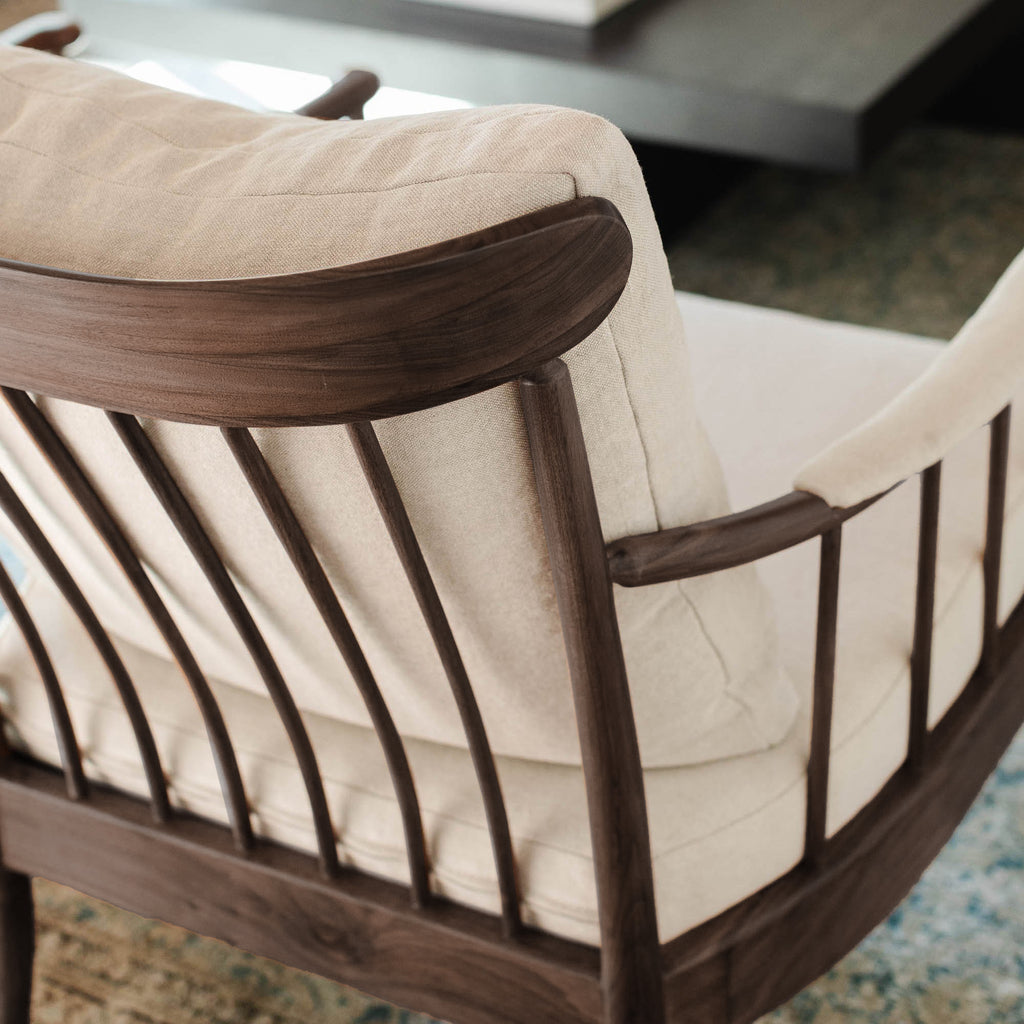 Close-up of a modern armchair with a dark brown wooden spindle backrest and thick cream-colored cushions, set on a patterned rug.