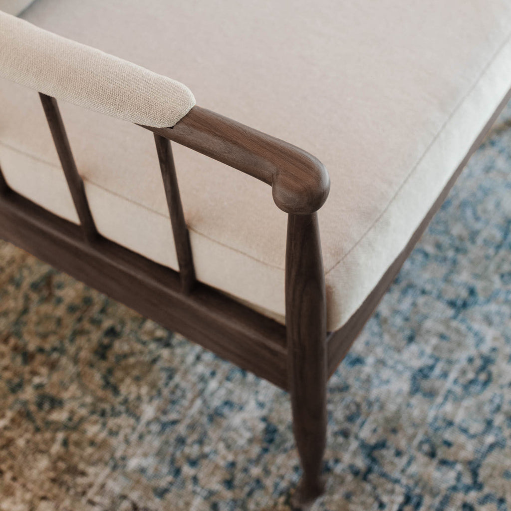 Close-up of a wooden lounge chair or chaise with a cream upholstered seat and backrest, shown resting on a blue and beige patterned area rug.
