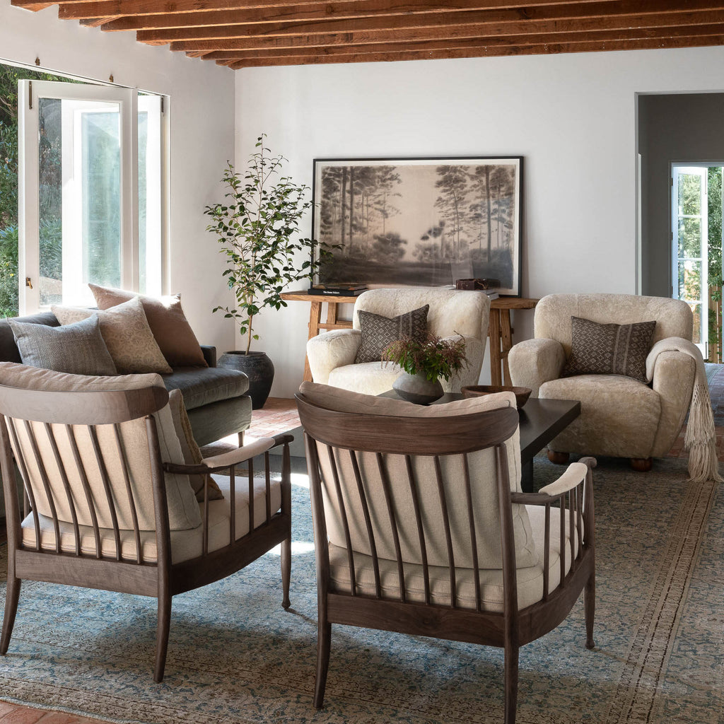 A styled living room featuring a coordinated set of armchairs and a sofa in neutral beige, cream, and brown tones, arranged around a dark wood coffee table on a patterned area rug. The room has white walls, exposed wooden ceiling beams, and a large framed landscape print.