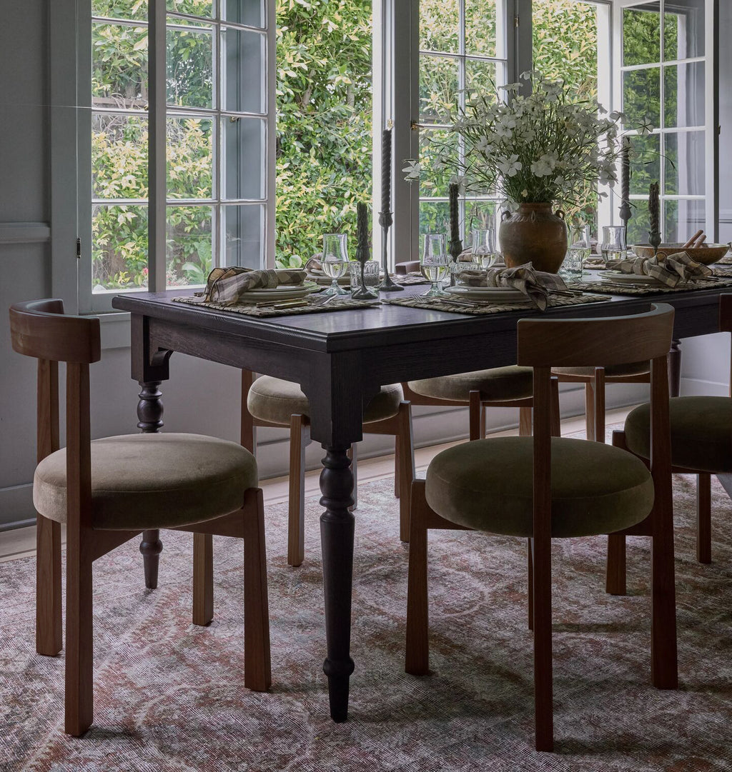 A dark wood dining table set with six upholstered wooden chairs in a bright dining room with large windows and greenery outside.