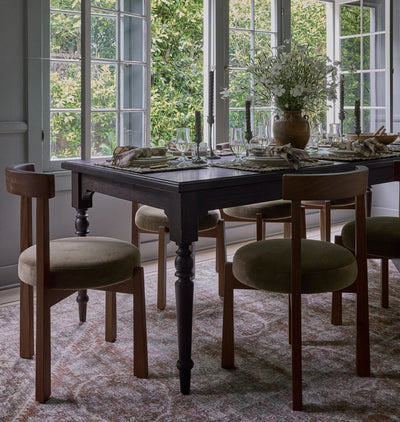 A dark wood dining table set with six upholstered wooden chairs in a bright dining room with large windows and greenery outside.
