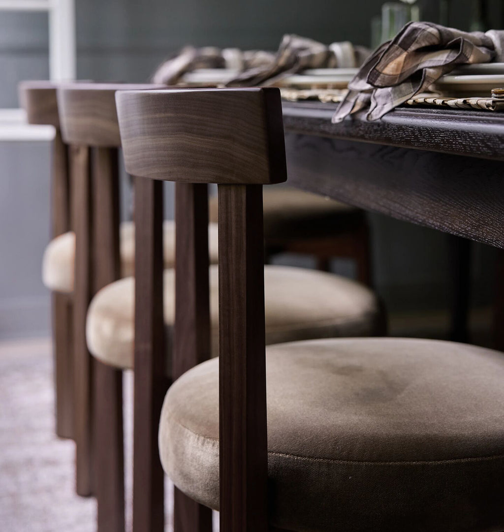A close-up of a dining table with upholstered bar stools. The stools feature dark wood backs and seats with beige fabric upholstery, set in a dimly lit dining room.
