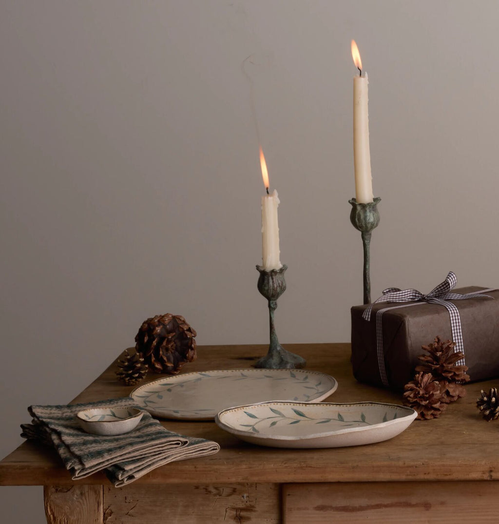A styled tabletop scene with two ceramic plates featuring a delicate green leaf pattern, set on a rustic wooden table alongside lit candles in metal holders, pinecones, a folded striped napkin, and a wrapped gift box against a neutral gray background.
