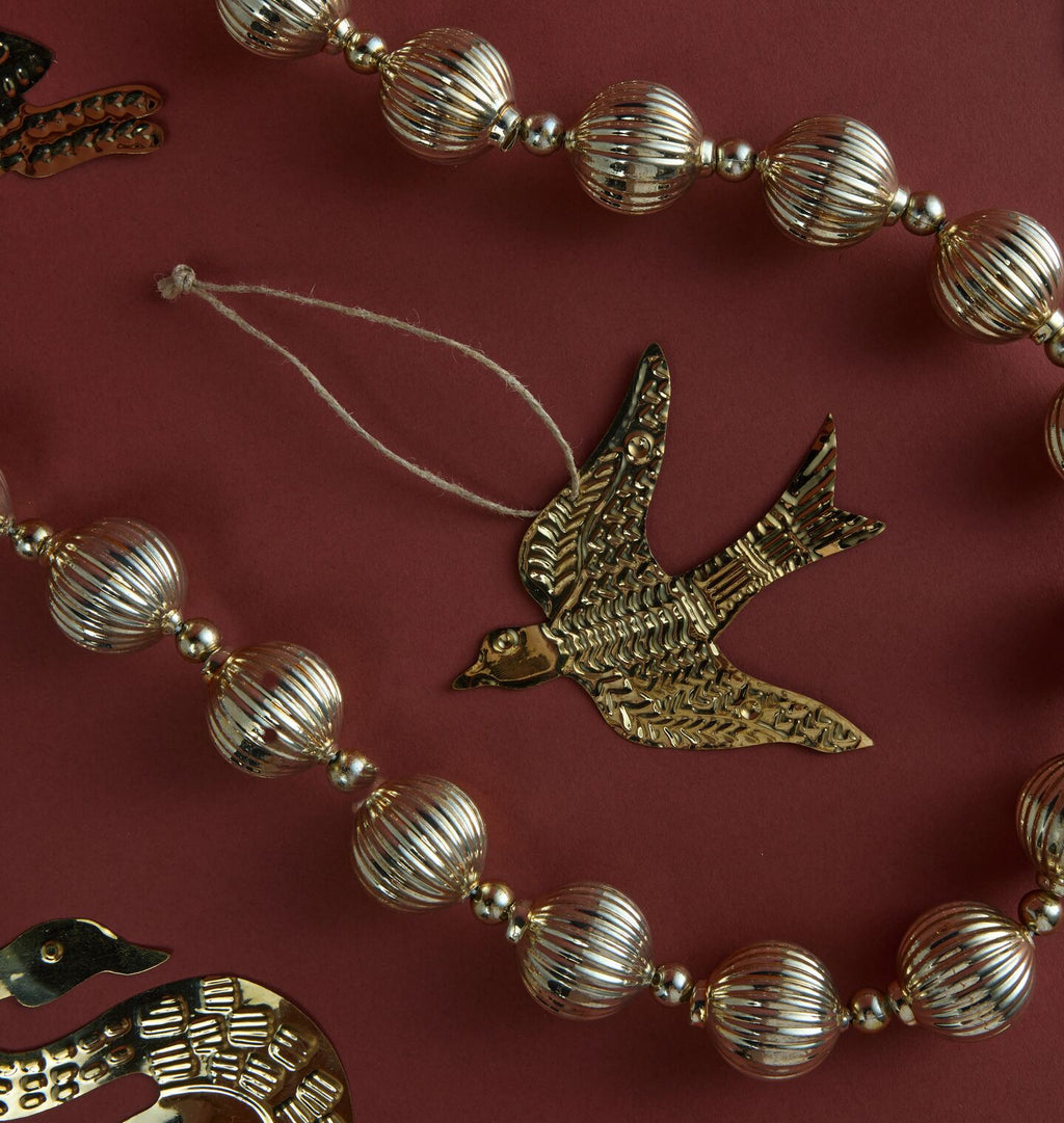 A close-up of a decorative garland featuring metallic gold-toned ribbed glass beads and a central bird-shaped ornament with a hanging string, arranged on a dark red background.