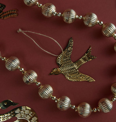 A close-up of a decorative garland featuring metallic gold-toned ribbed glass beads and a central bird-shaped ornament with a hanging string, arranged on a dark red background.
