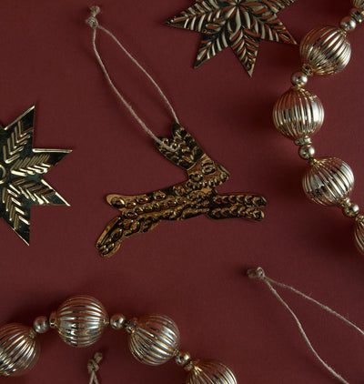 A styled flat-lay of metallic Christmas ornaments on a dark burgundy background, featuring a central gold reindeer rabbit and ribbed gold and silver bead garlands.