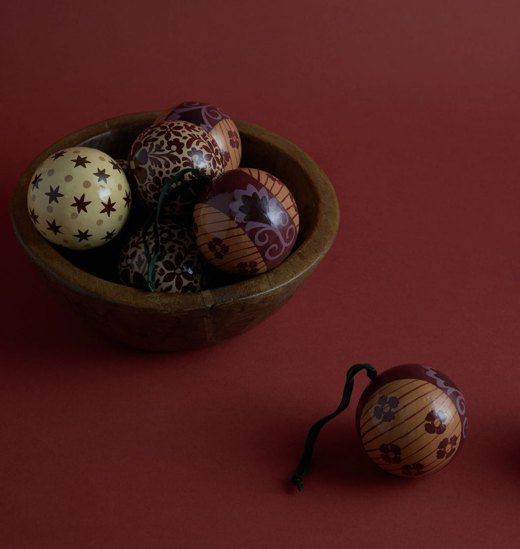 A set of hand-painted paper mache ball ornaments displayed in a dark wooden bowl, with one ornament resting outside the bowl on a solid dark red background.
