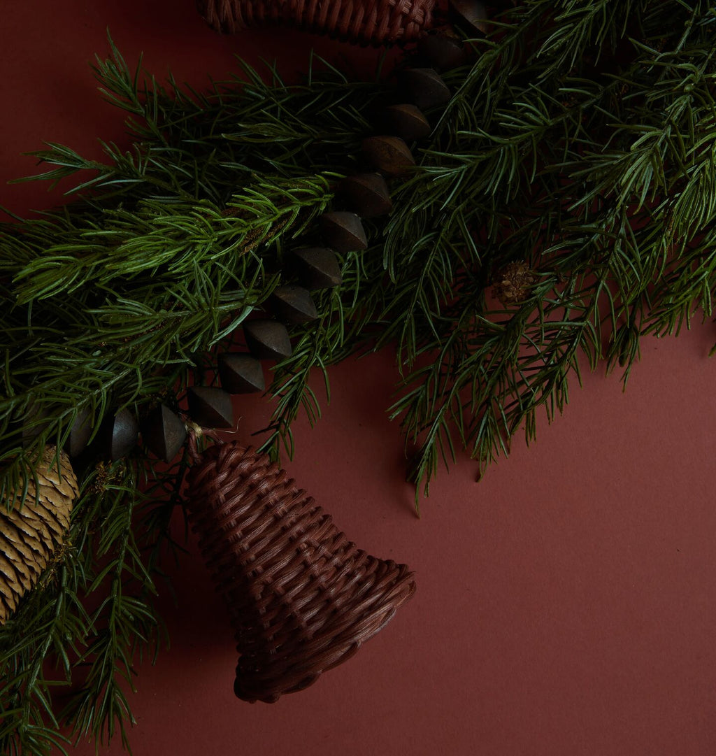 A dark brown woven wicker Christmas wreath with pinecones and a hanging wicker bell, arranged on a dark red background with green pine branches.