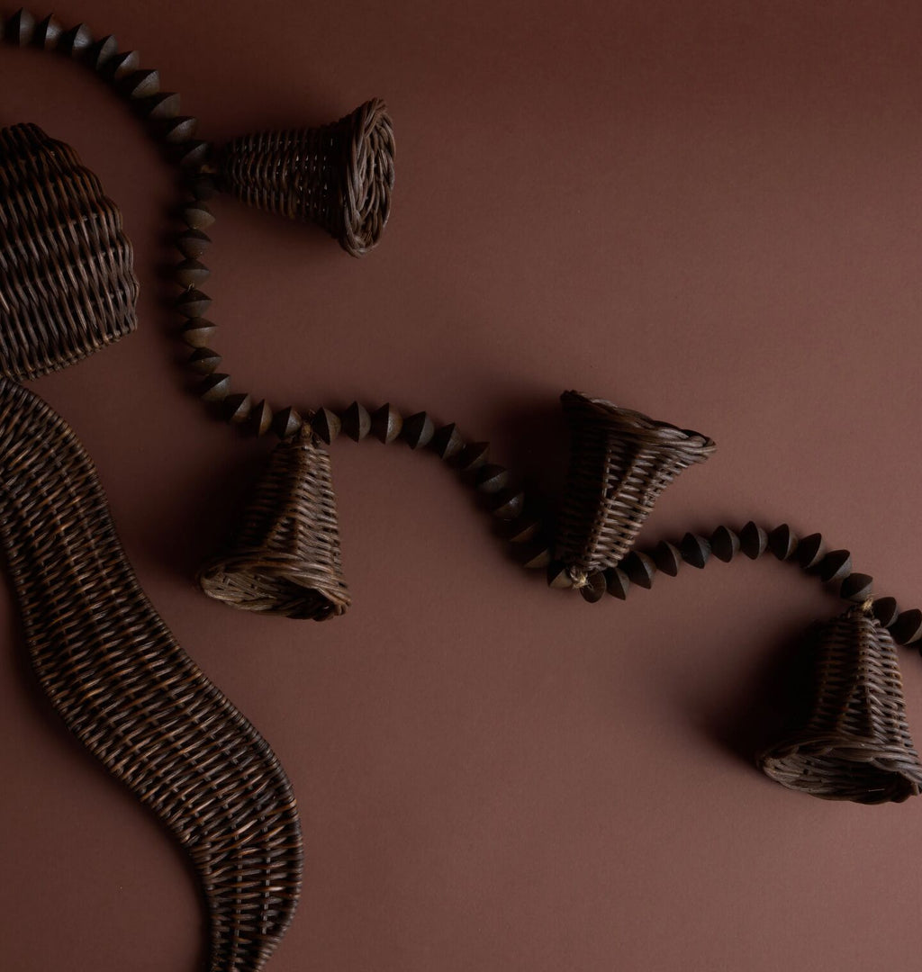 Dark brown woven wicker garland with carved beads and bell-shaped ornaments, arranged on a solid dark brown background.