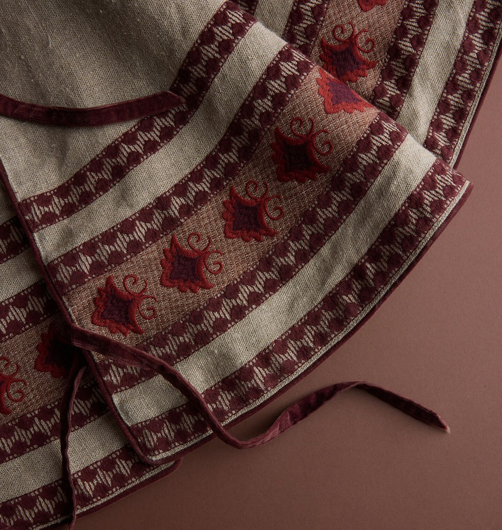 A close up of a tree skirt with red and brown embroidery on a beige fabric with red velvet bow ties on a brown background