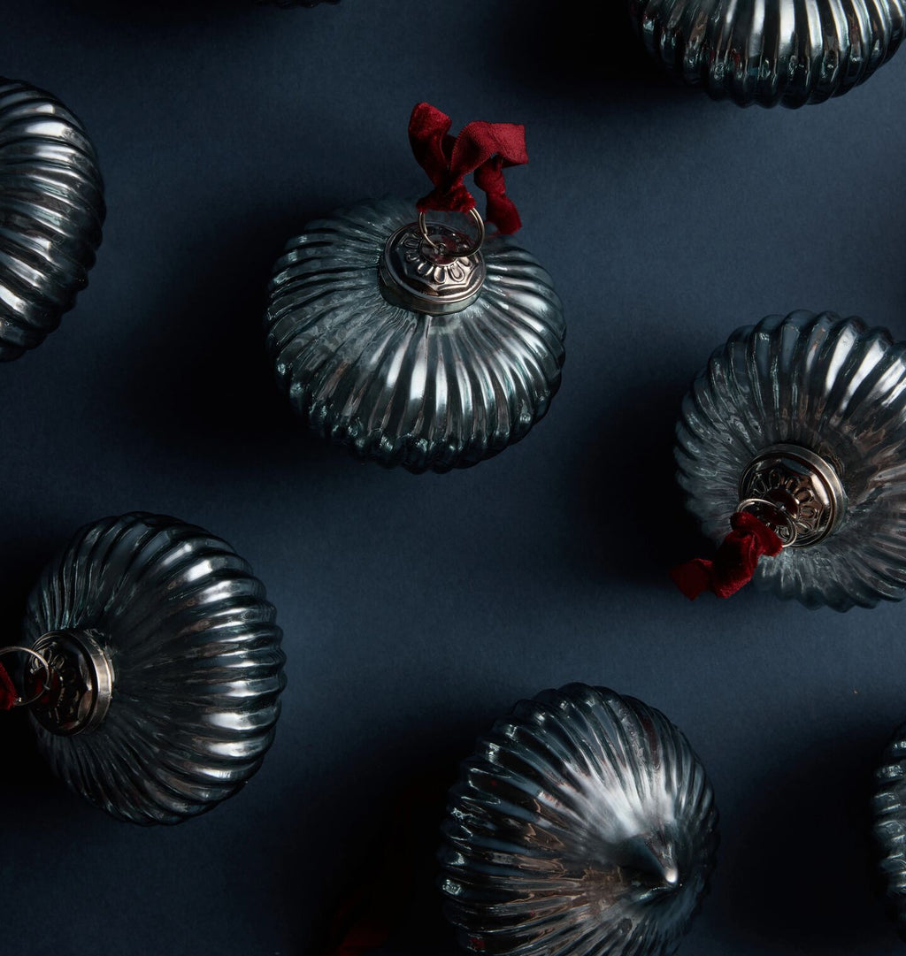 Several ribbed glass Christmas ornaments in a smoky gray color, arranged on a dark navy background. One ornament near the top center has a small red ribbon loop and a silver metal cap.