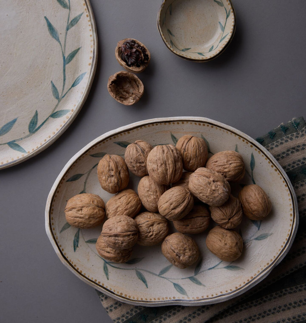An oval ceramic serving platter filled with whole walnuts, styled on a dark gray surface with other matching plates and bowls. The platter has a cream glaze, a delicate green leaf pattern, and a gold rim.