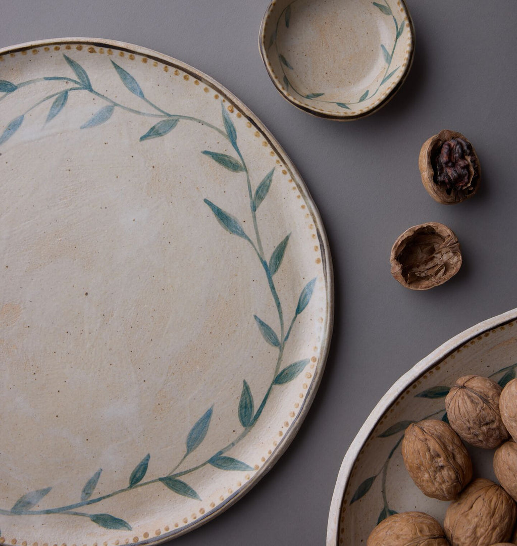 A ceramic serving platter with a leafy vine border, shown alongside matching bowls and walnuts on a dark gray surface.