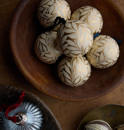 A group of hand-painted wooden ornaments, decorated with a gold leaf pattern on a cream background, arranged in a rustic wooden bowl.