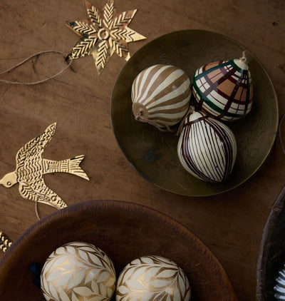 Three hand-painted Christmas ornaments with traditional patterns, displayed in rustic wooden bowls alongside wooden bird and star ornaments on a dark wood background.