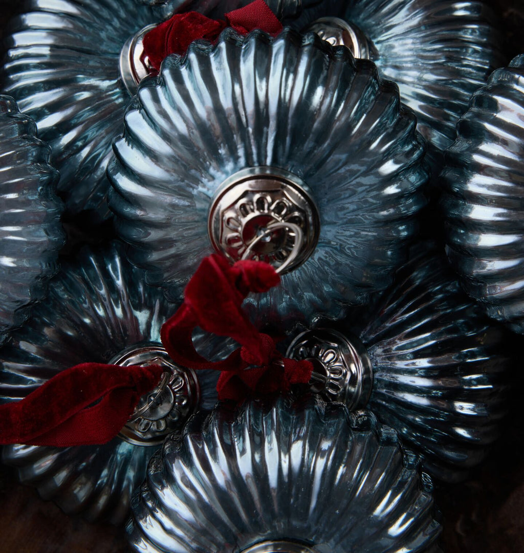 A close-up of several ribbed glass ornaments in a smoky blue-gray color, each with a silver-toned metal cap and a red velvet ribbon, tightly packed together.