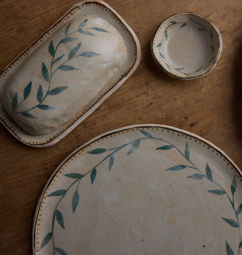 A rustic ceramic tableware set on a wooden surface, featuring a large oval plate, a rectangular butter dish, and a small bowl, all decorated with a blue-green leafy vine pattern and a gold beaded rim.