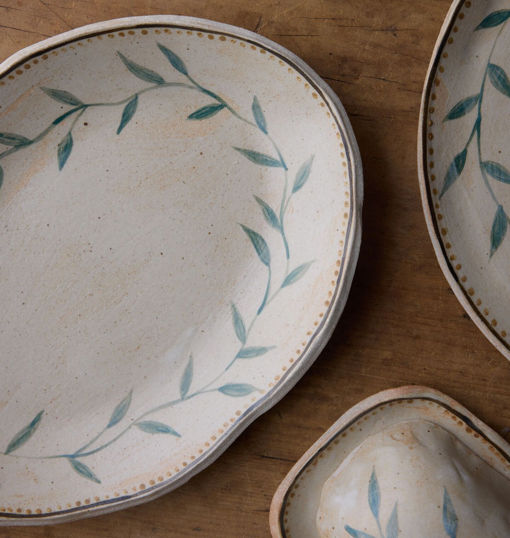 A close-up of a ceramic serving platter with an organic rim, decorated with a green leafy vine border and a gold dotted accent line, set on a wooden surface.