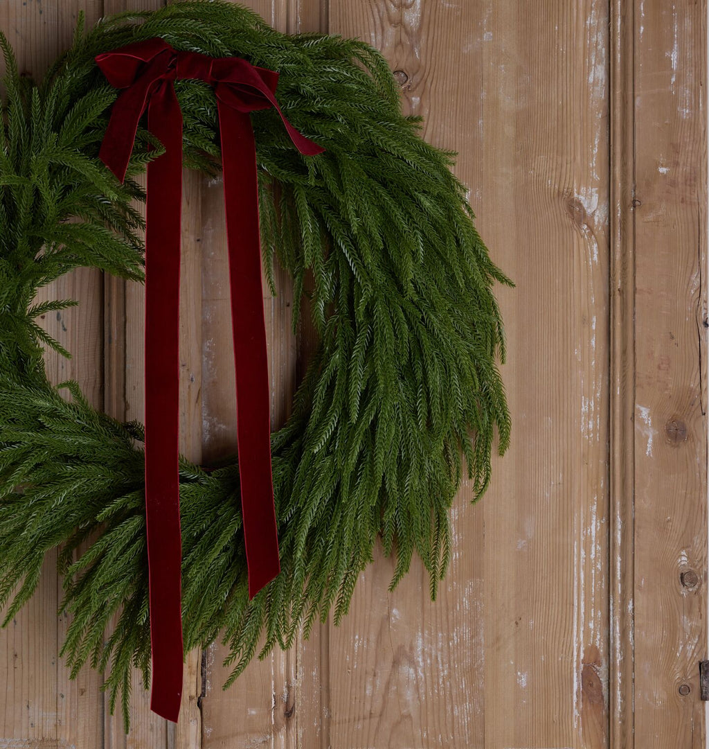 A green pine wreath with a long dark red velvet ribbon bow, hanging against a rustic wooden door.