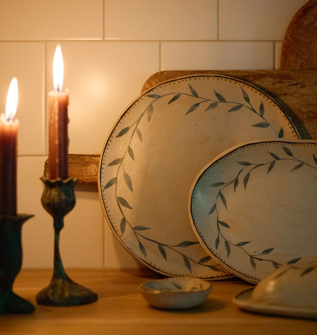 Two round ceramic plates with a blue-green leafy vine border and dark rim detailing, styled on a wooden surface next to lit candles and a small bowl.
