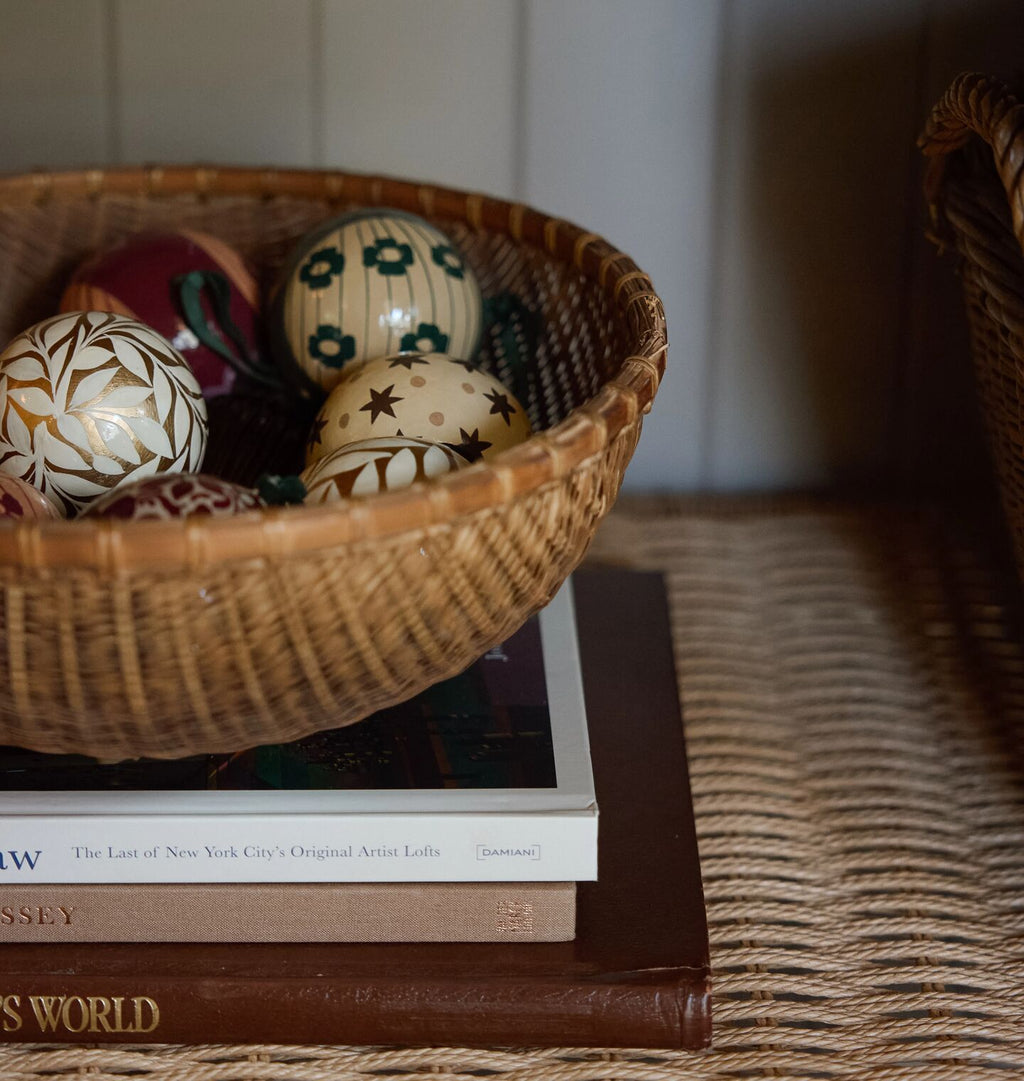 A woven wicker basket filled with decorative patterned eggs, styled on top of stacked books in a warm indoor setting.
