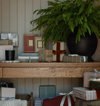 A styled holiday gift scene with wrapped presents, a large black vase holding a green pine branch arrangement, and framed art displayed on a wooden console against a paneled wall.