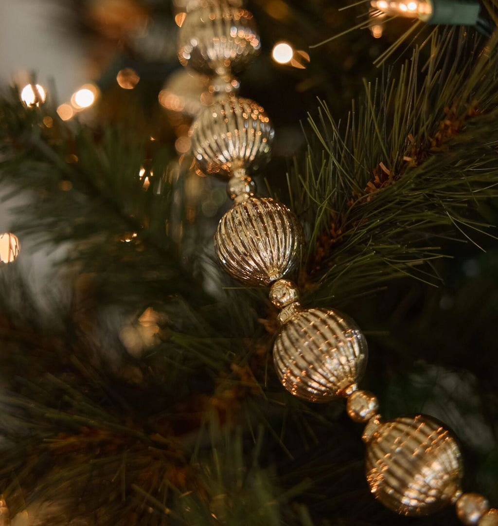 A close-up of a gold beaded Christmas garland with ribbed, textured spherical ornaments draped over dark green pine tree branches with warm string lights in the background.
