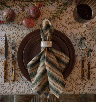 An overhead shot of a set table with reddish brown plates, a patterned table cloth. bistro silverware set and a striped embroidered napkin in beige and muted green with a ceramic napkin ring