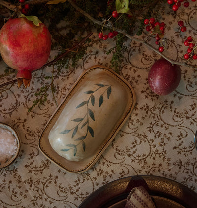 A warm christmas table setting scene featuring a patterned tablecloth and a pomegranate and plum arrangement featuring a handmade ceramic butter dish with a vine motif on top