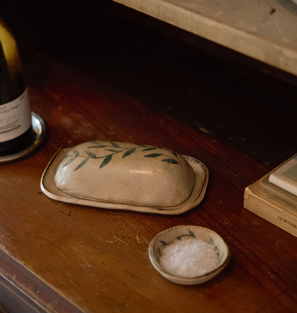 A rustic ceramic butter dish with a matching small bowl of coarse salt, styled on a dark wooden table next to a wine bottle and books.