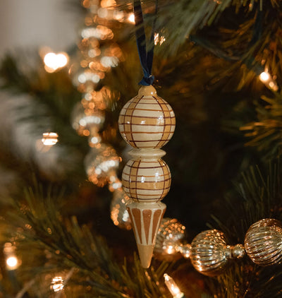 A decorative turned wood Christmas ornament with a painted plaid pattern hanging on a lit Christmas tree with green needles and warm bokeh lights in the background.