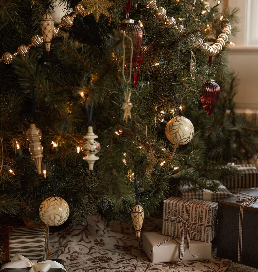 A decorated Christmas tree in a home setting, adorned with warm white and gold ornaments, string lights, and ribbons, with wrapped gift boxes placed underneath on a patterned rug.