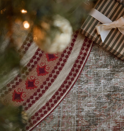 A close-up of a Christmas tree skirt with a red and beige embroidered geometric pattern, styled under a Christmas tree with a wrapped gift box and warm bokeh lights.