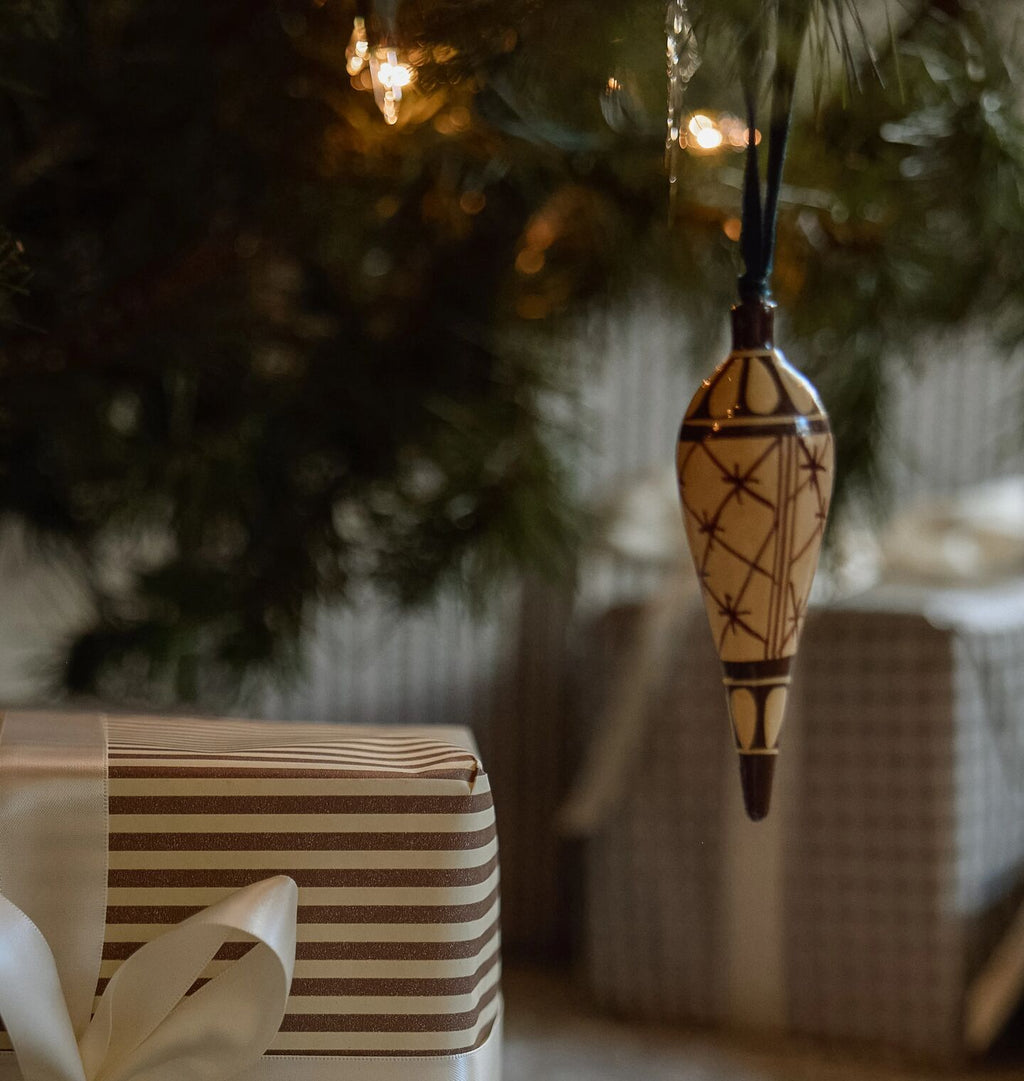 A decorative Christmas ornament hangs from a tree branch above wrapped gifts. The ornament is a teardrop shape with a cream background and dark brown geometric star patterns, suspended by a dark ribbon.