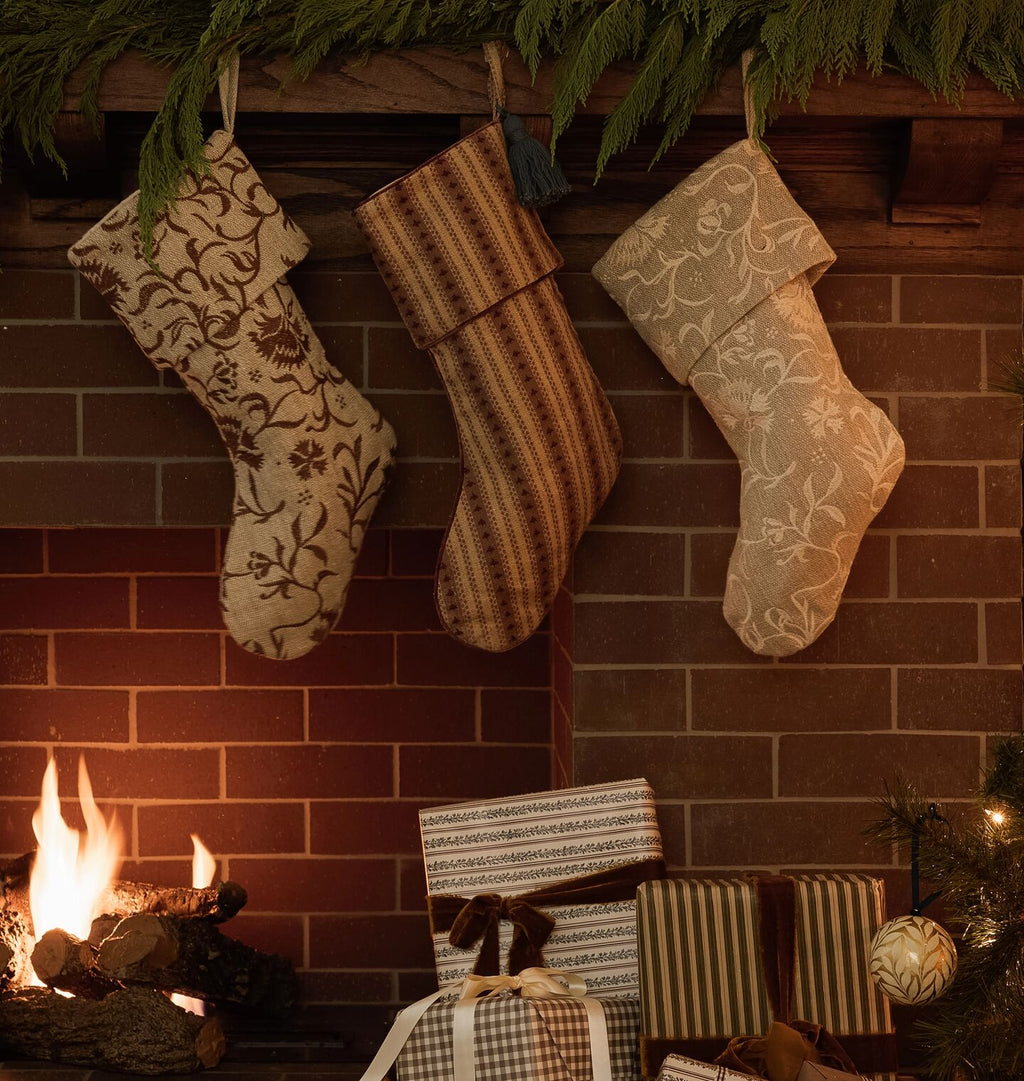 Three decorative Christmas stockings hang on a brick fireplace mantel, with a burning fire below and wrapped gifts at the base. The stockings feature floral and striped patterns in neutral tones.