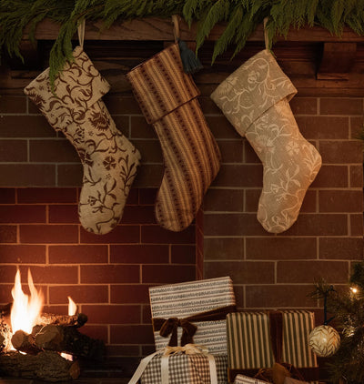 Three decorative Christmas stockings hang on a brick fireplace mantel, with a burning fire below and wrapped gifts at the base. The stockings feature floral and striped patterns in neutral tones.
