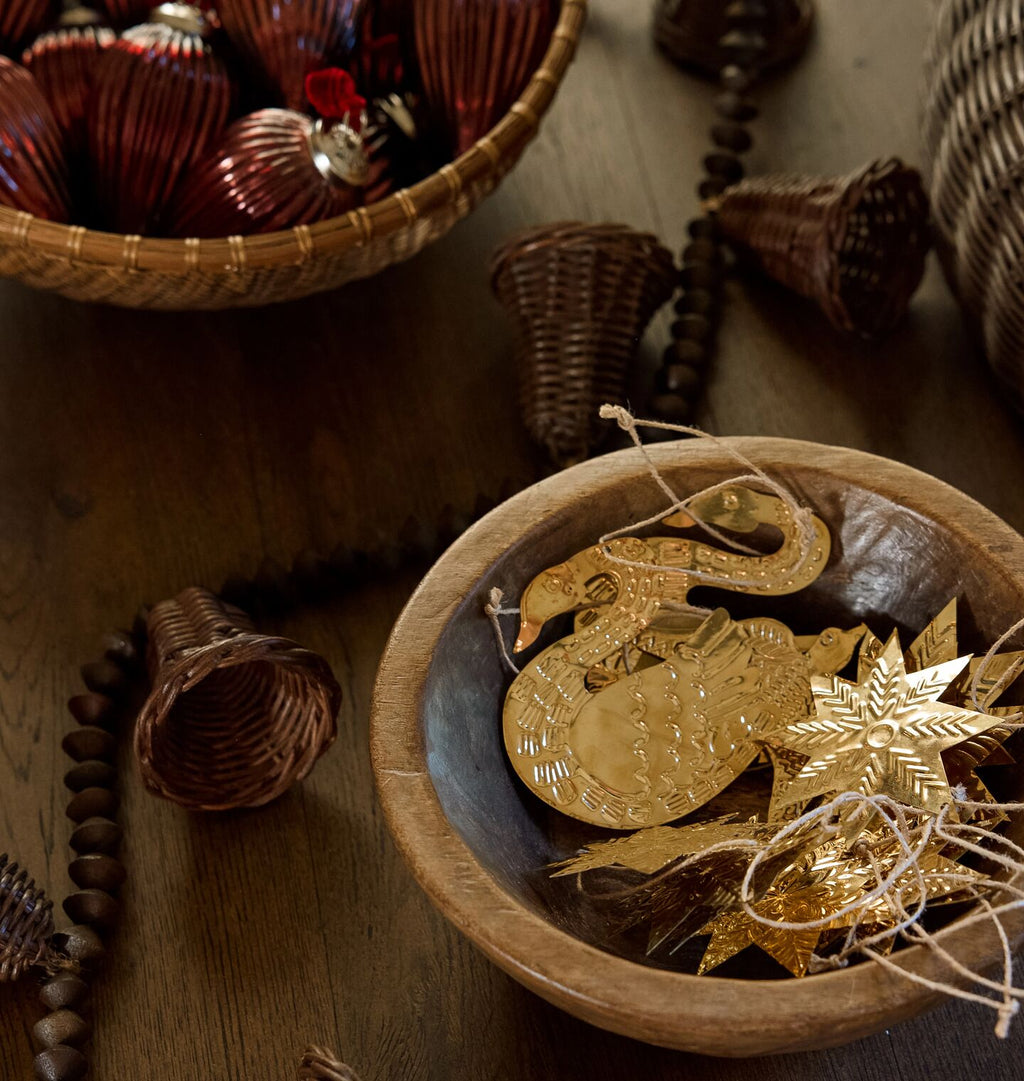 Decorative gold-toned Christmas ornaments, including a swan and a star, displayed in a wooden bowl on a rustic wooden table, with a basket of red and gold ornaments in the background.