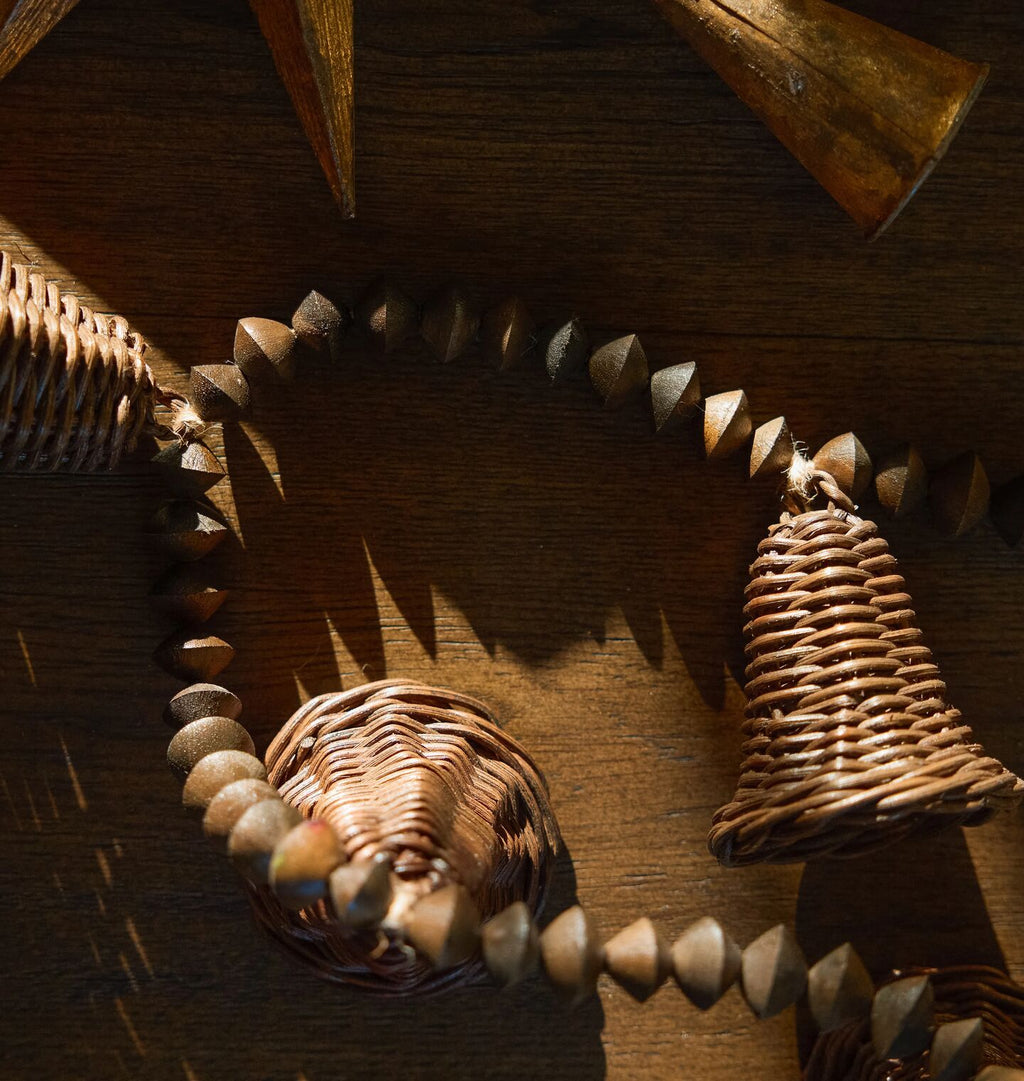 A rustic collection of handcrafted wooden and wicker garland arranged on a dark wood surface under dramatic, direct sunlight. The garland includes wood beads and a wicker bell.