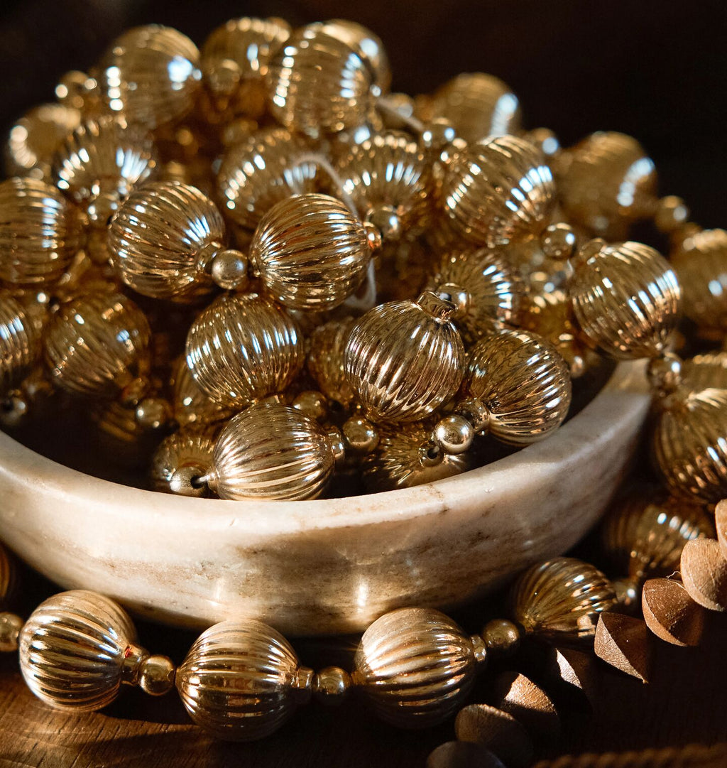 A close-up of metallic gold-toned beaded garland, including ribbed round beads and small spacer beads, arranged around and inside a shallow white dish on a dark wooden surface.
