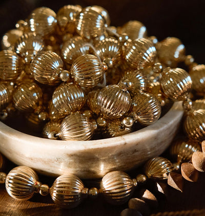A close-up of metallic gold-toned beaded garland, including ribbed round beads and small spacer beads, arranged around and inside a shallow white dish on a dark wooden surface.