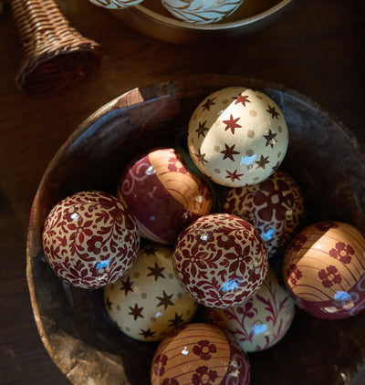 Decorative hand painted paper machet ornaments with floral and star patterns, arranged in a dark wooden bowl on a dark wooden table.