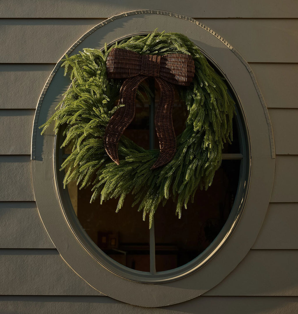 A green pine wreath with a wicker dark brown ribbon bow, hanging against a oval window.