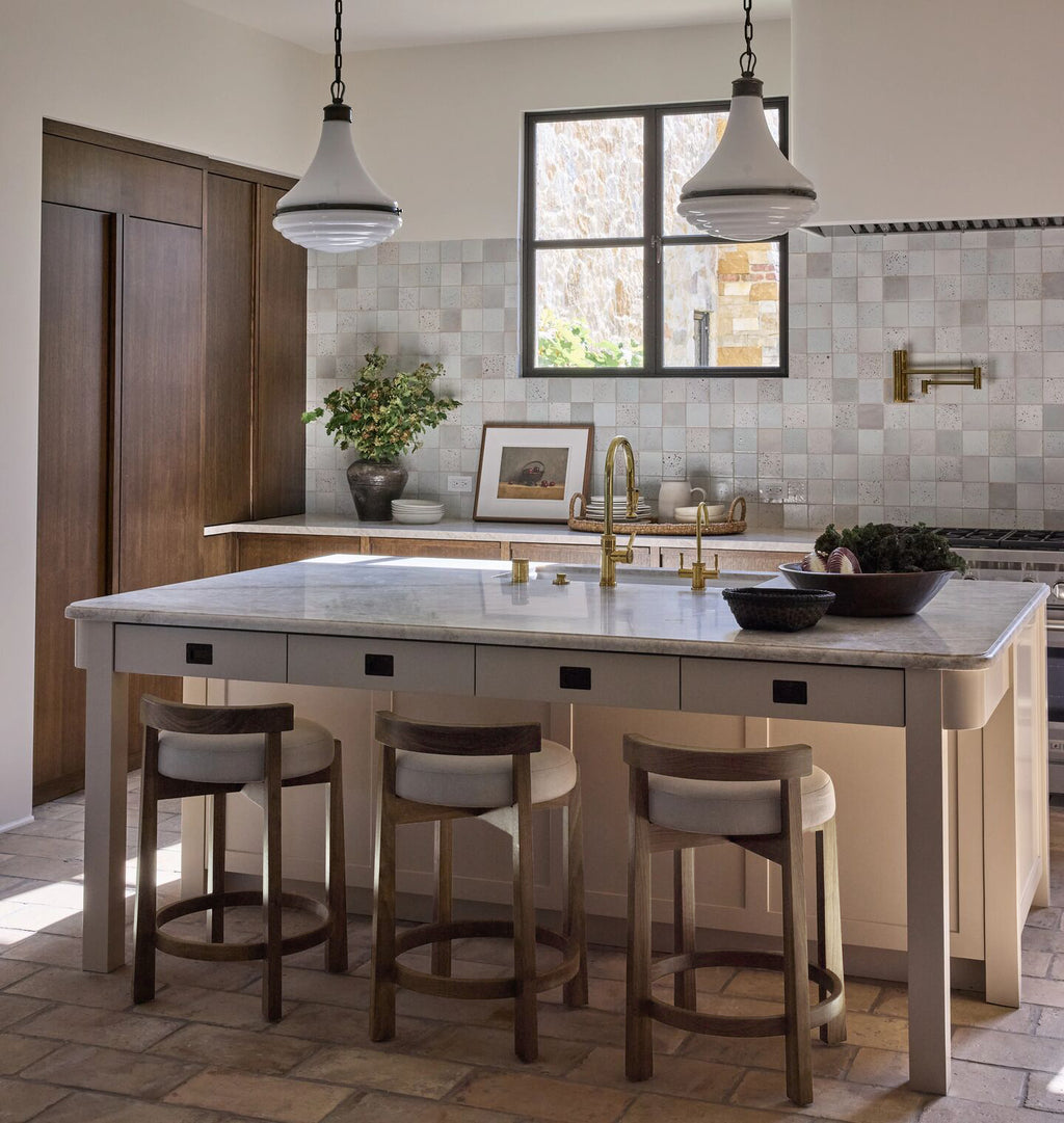 A modern kitchen island with a light stone countertop, white drawers, and four wooden bar stools with light cushions, set in a bright kitchen with tiled backsplash and pendant lights.
