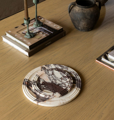 Two round coasters with a dark brown and cream marble pattern arranged on a light wood tabletop, styled with books, candlesticks, and a ceramic pot.