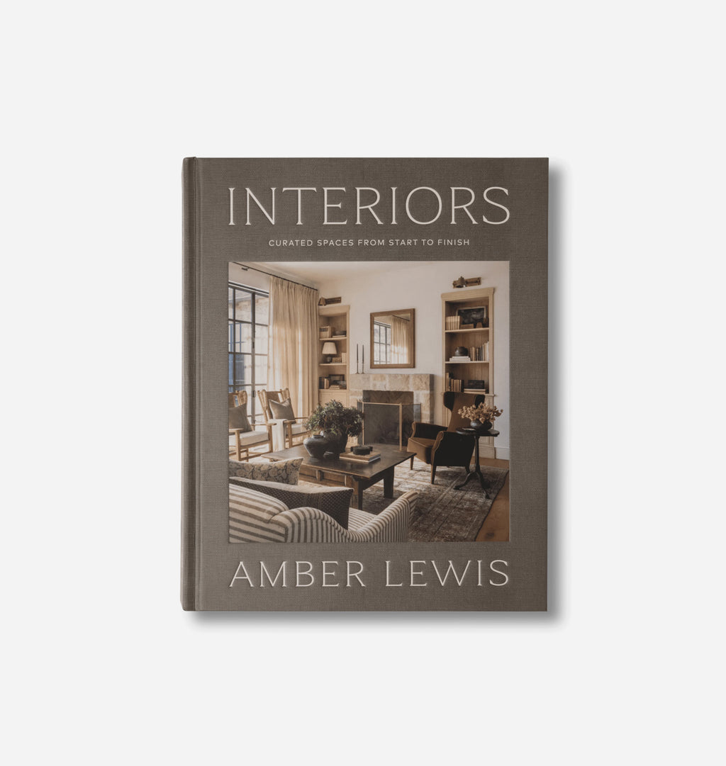 The book 'Interiors: Curated Spaces from Start to Finish' by Amber Lewis, featuring a dark taupe-gray cover with a central photograph of a cozy living room, set against a light gray background.