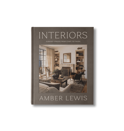 A hardcover book titled 'Interiors' by Amber Lewis, featuring a warm-toned interior photograph on the cover, set against a clean off-white background.